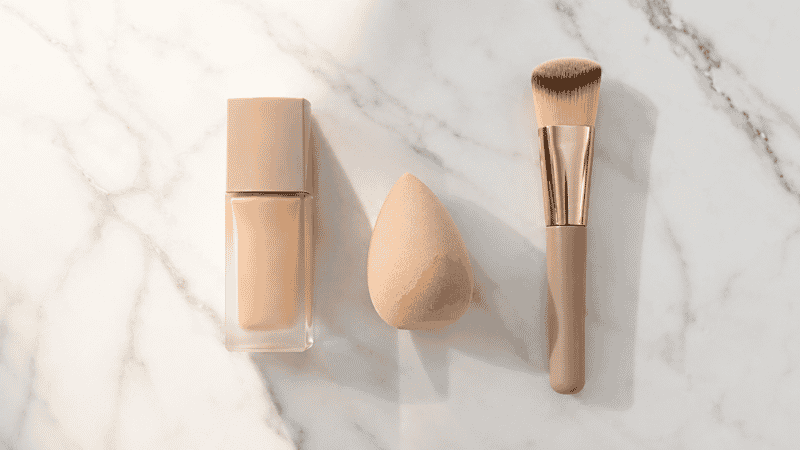 Foundation bottle, beauty sponge and brush arranged on white marble for a beginner makeup guide