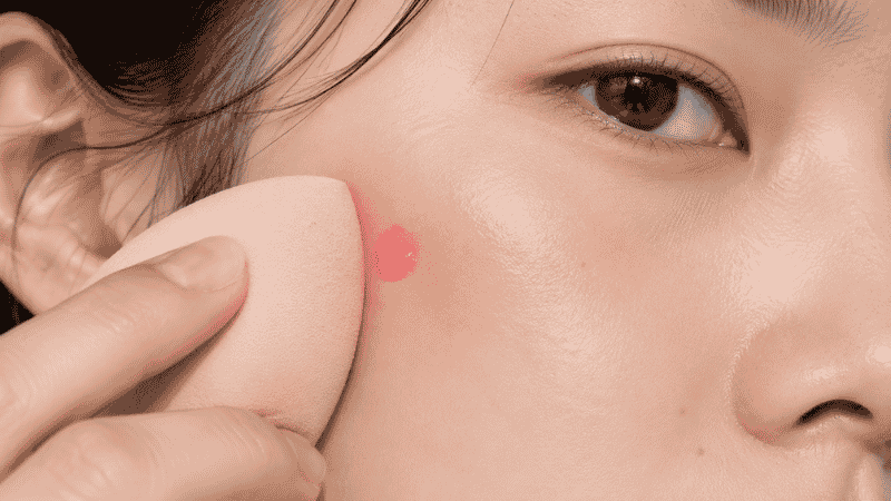 One small drop of liquid blush being blended onto skin with a beauty sponge