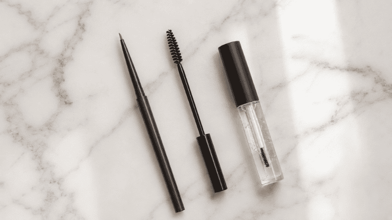 Eyebrow pencil, spoolie brush and brow gel arranged on white surface for a beginner eyebrow tutorial