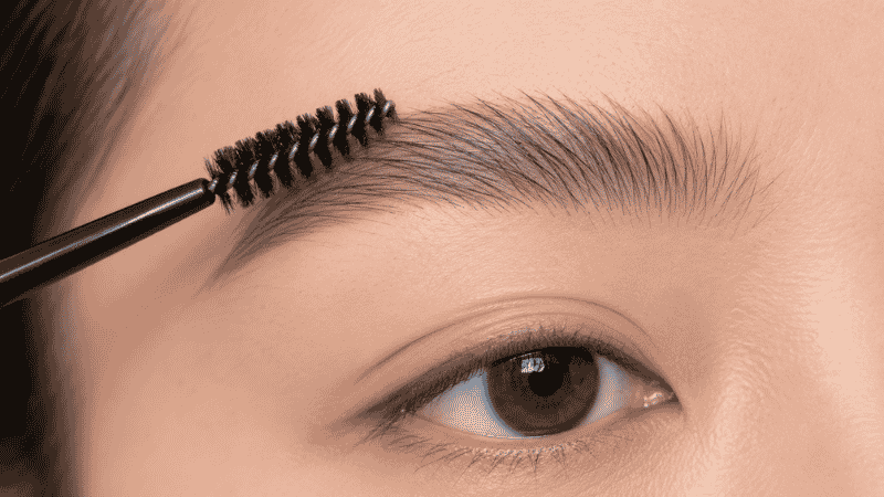 Spoolie brush combing eyebrow hairs upward before filling to reveal natural brow shape