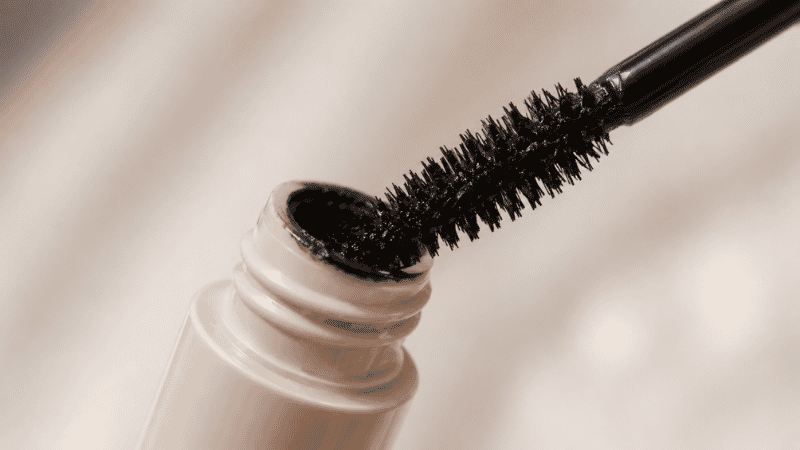 Mascara wand being wiped against the tube opening to remove excess product before application