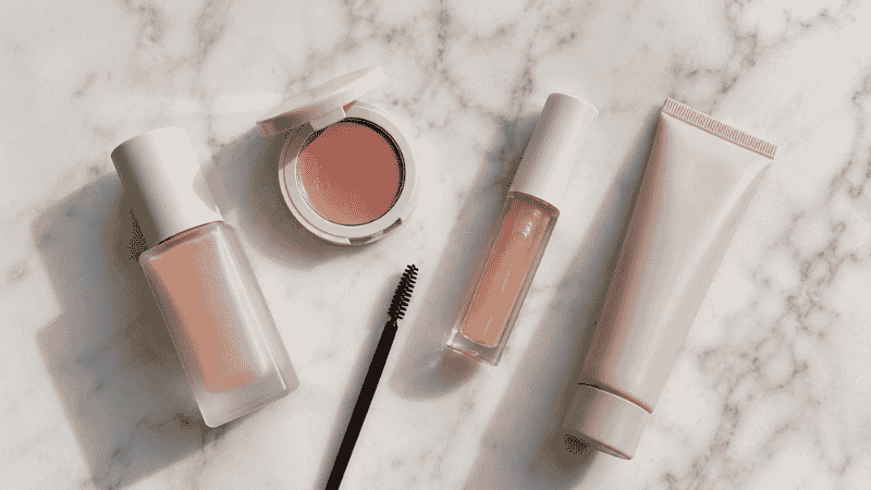 Minimal makeup products including skin tint, cream blush and lip gloss arranged on white surface for a natural beauty routine