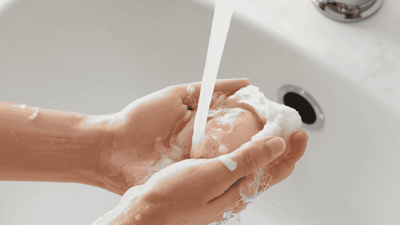 A person washing a pink beauty blender sponge under running water to remove bacteria and old sebum, preventing breakouts for oily and acne-prone skin.