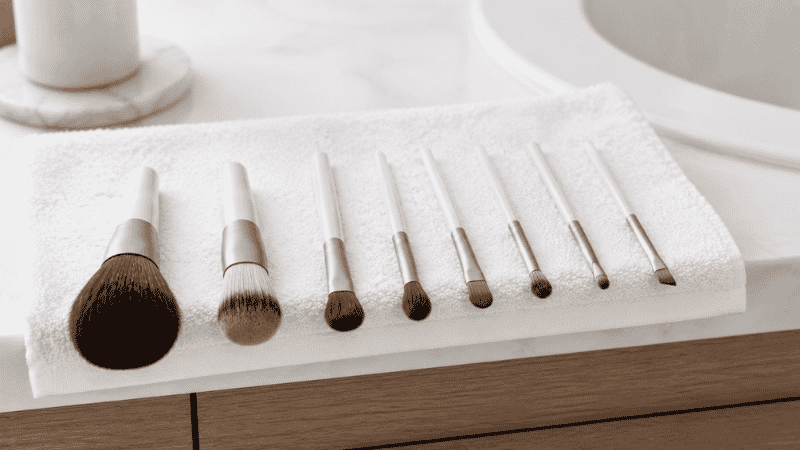 Makeup brushes laid flat on a white towel with bristles hanging off the edge of a counter to dry correctly